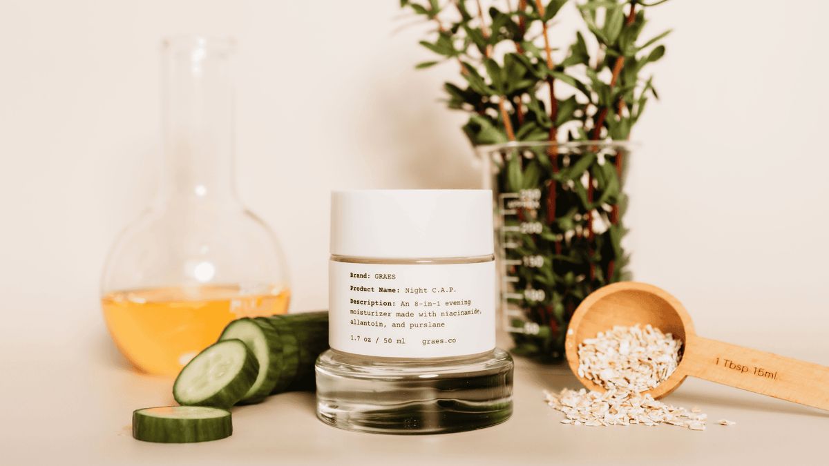 GRAES Night C.A.P. glass jar with label, sliced cucumber, wooden scoop with oatmeal, flask of jojoba oil, and purslane on a light surface.